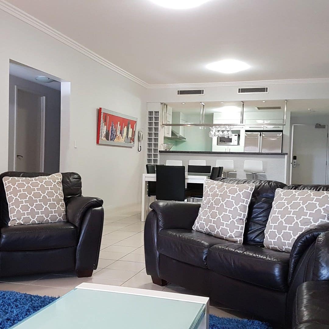 Open living room and kitchen, beige tile, white walls, black sofas, tan throw pillows, blue area rug, glass coffee table.