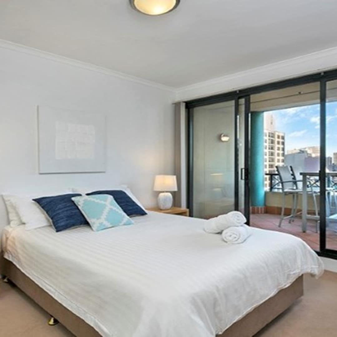 A bedroom with beige carpet, a queen size bed, white and blue bedding, and sliding glass doors to a balcony with city views.