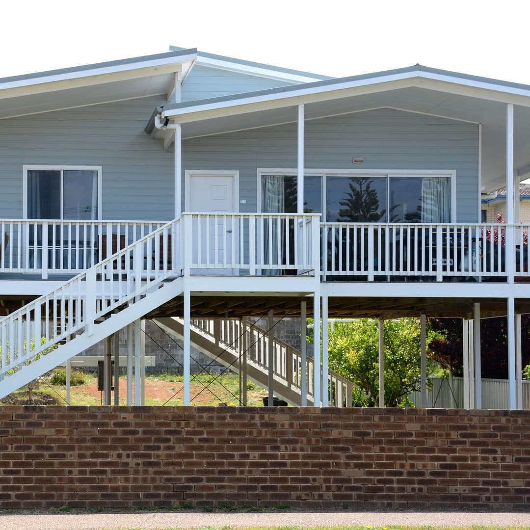 Exterior of a raised home with light blue siding, wrap-around porch, wide windows, front yard enclosed by short brick wall.