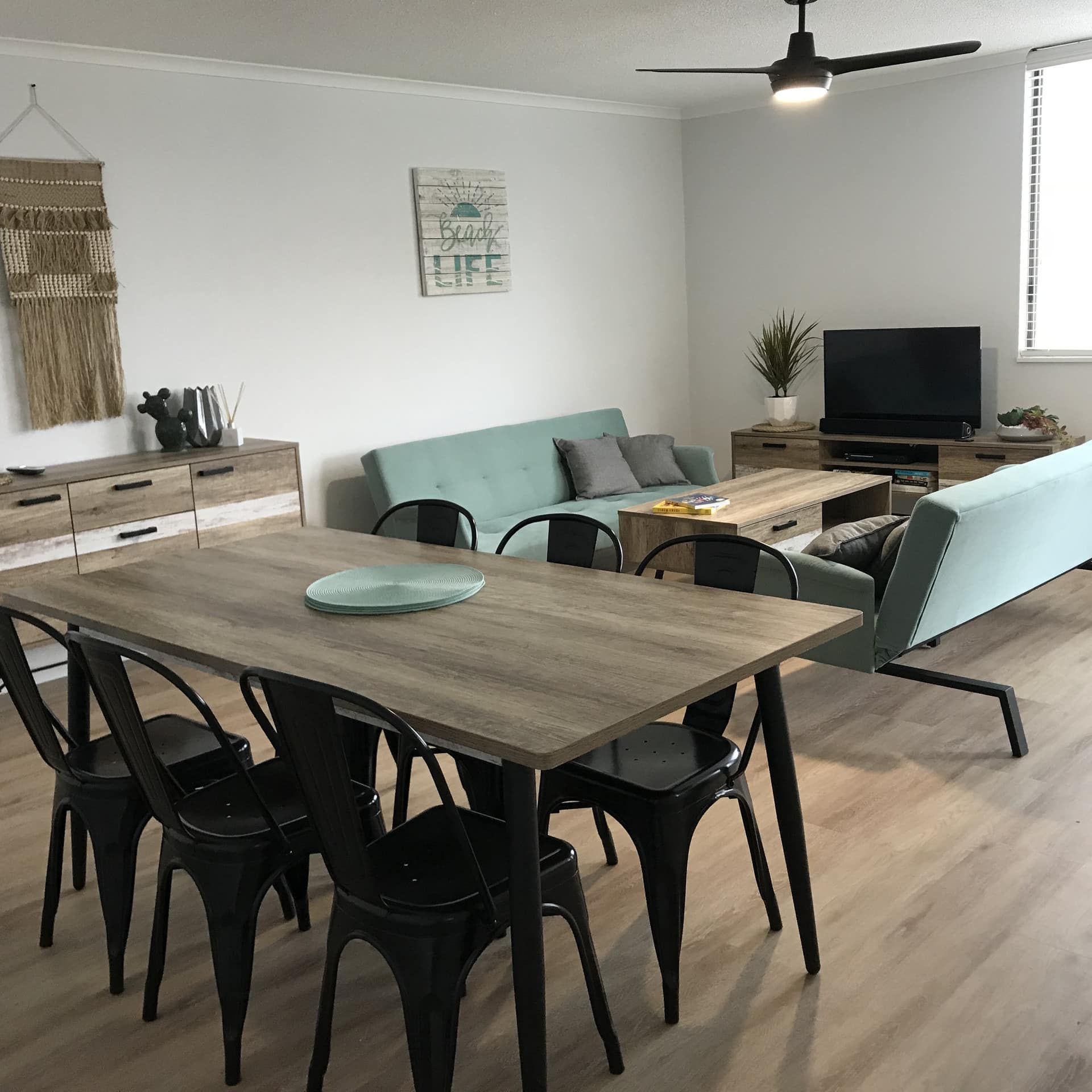 Modern, open dining and living room with hardwood floors, teal sofas, wood dining table, black chairs, woven wall hanging.