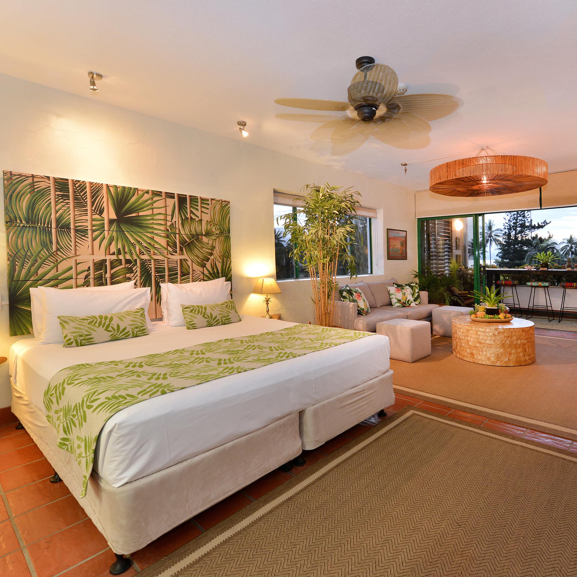 Tropical-themed open bedroom with a king size bed, seating area, and glass sliding doors to attached balcony.