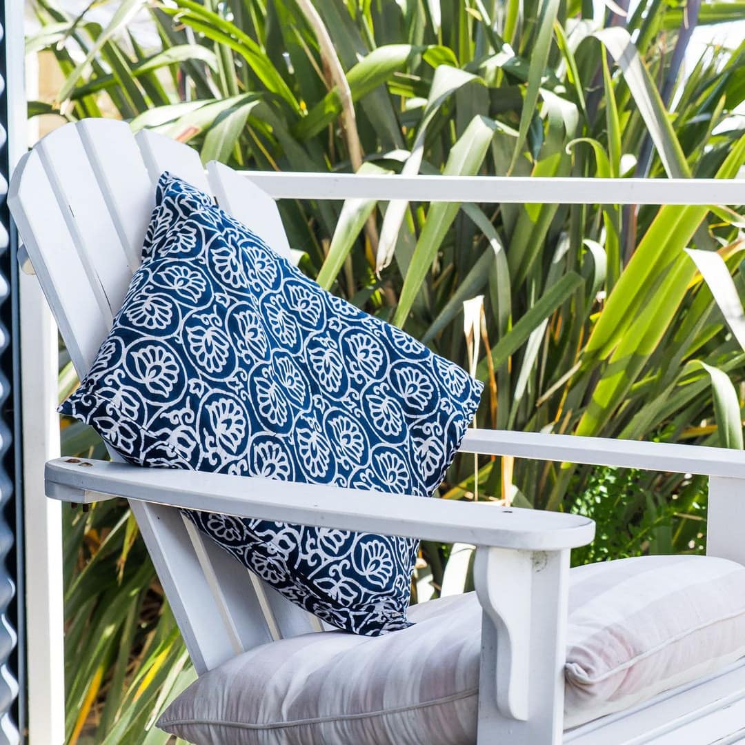A white, wooden lounge chair with blue and tan decorative pillows sits on a deck surrounded by palm trees.