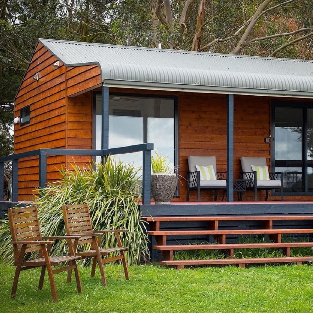Wood panelled cottage with grey roof, covered porch, grey lounge chairs, potted plants, and a lush lawn.