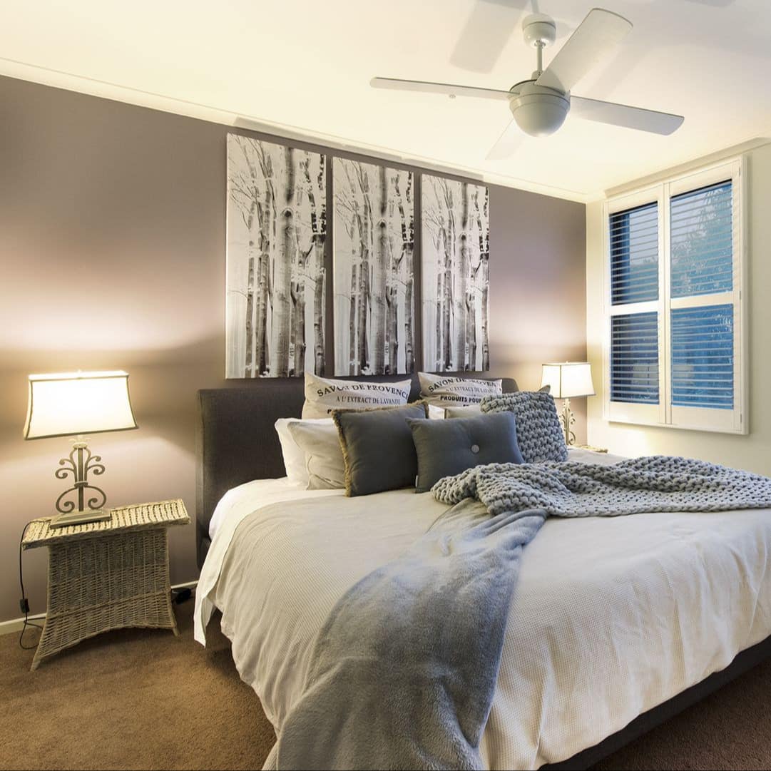 Spacious bedroom with grey accent wall, king-size bed, grey and white bedding, wicker table and black and white art.