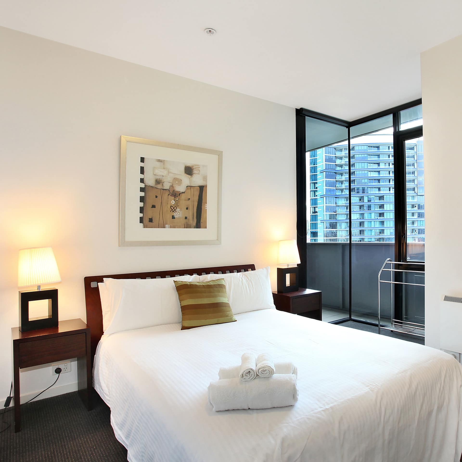 A bedroom with grey carpet, white walls, a queen size bed, white bedding, two wooden bedside tables, corner windows.