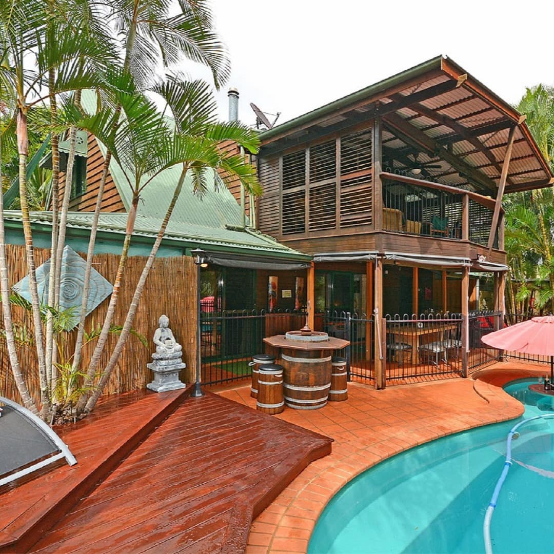 Bali-style 2-storey home with covered porches, pool with red tile patio and deck surrounded by lush vegetation.