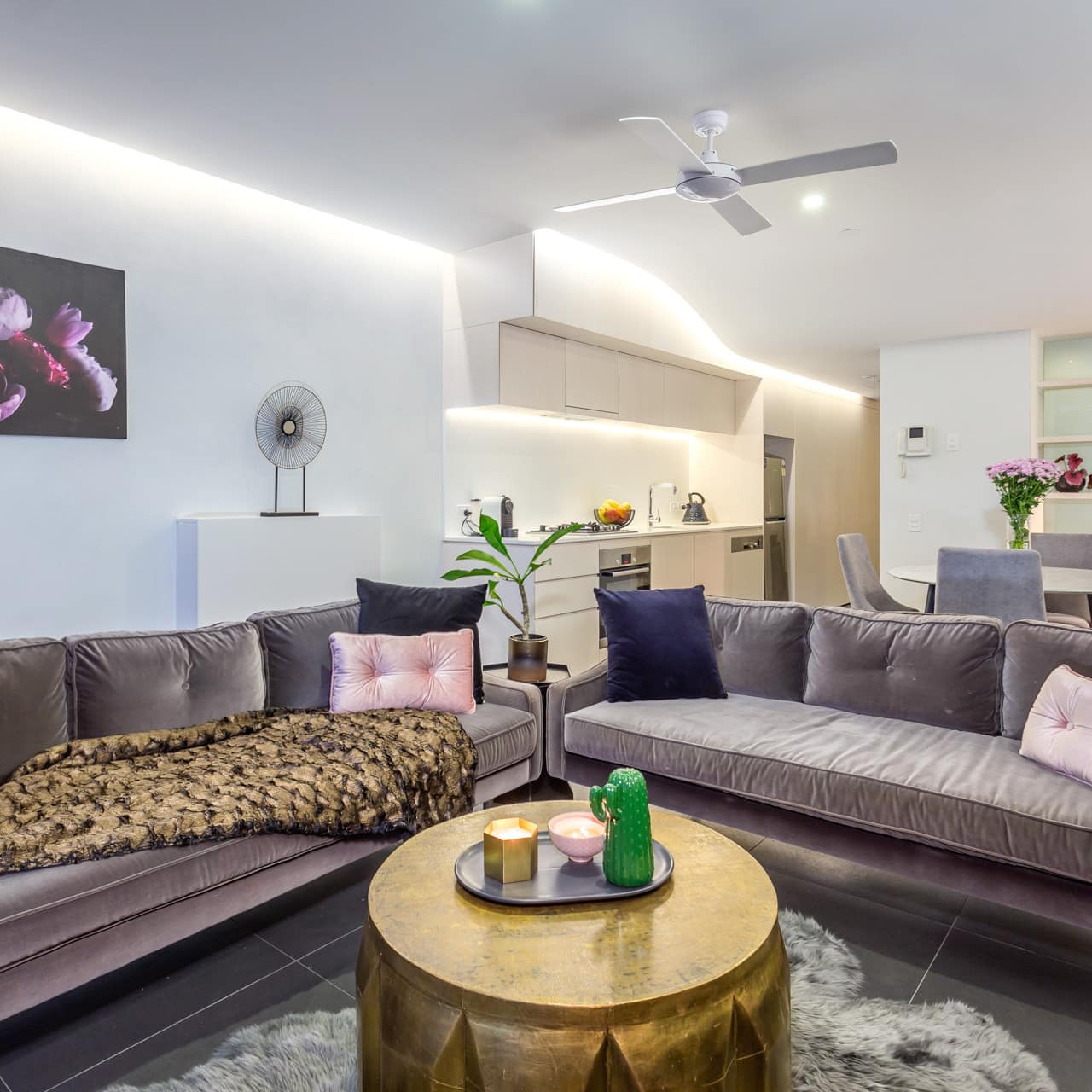 Open living room with grey tile floors, white walls, grey velvet sofas, gold coffee table, modern kitchen, and dining area.