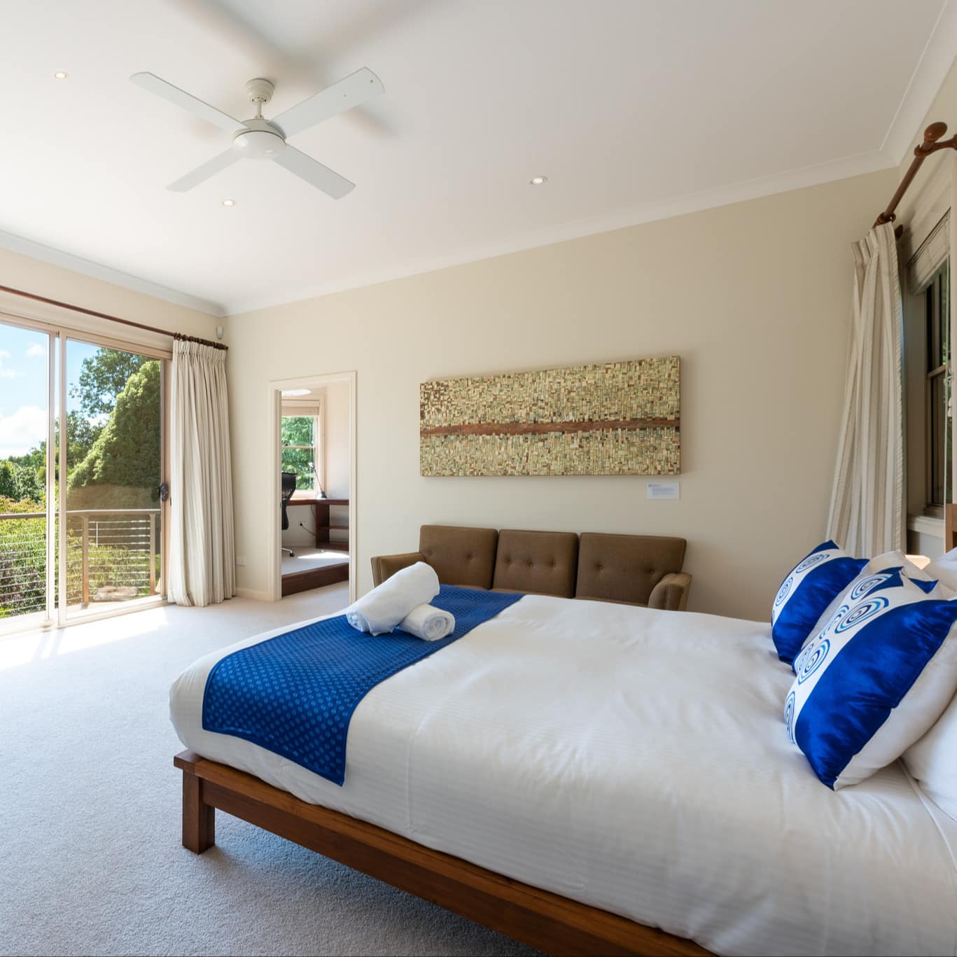 Master bedroom with king-sized bed, white linens, royal blue pillows, day bed and sliding glass windows with treetop view.