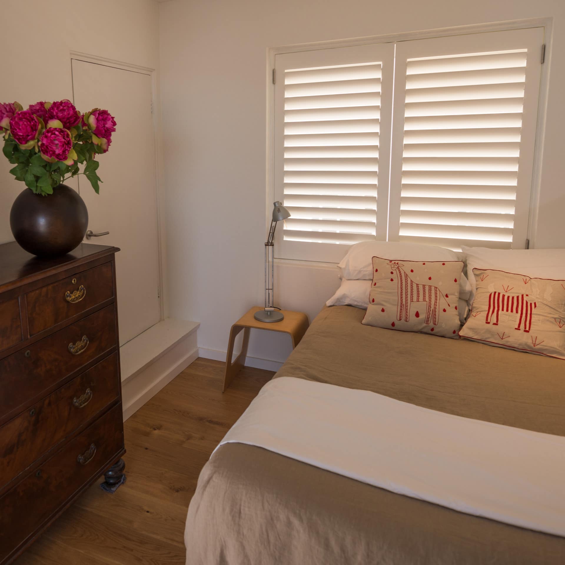 Bedroom with hardwood floors, white walls, queen size bed with tan bedding, and a wooden antique dress.