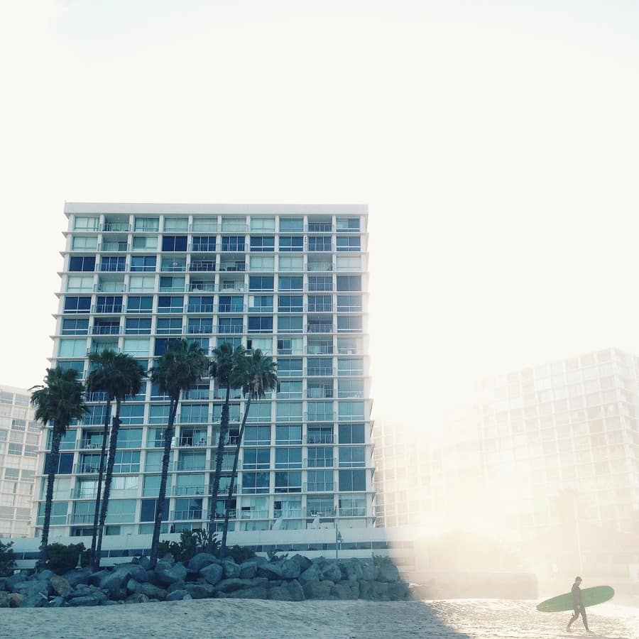 15-storey beachfront apartment building with palms and surfer walking with surfboard in sunlight
