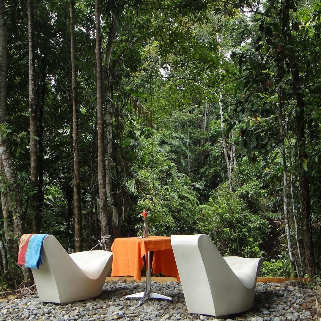 Two white, modern lounge chairs and a small metal table sit on a gravel patio surrounded by thick, lush rainforest.