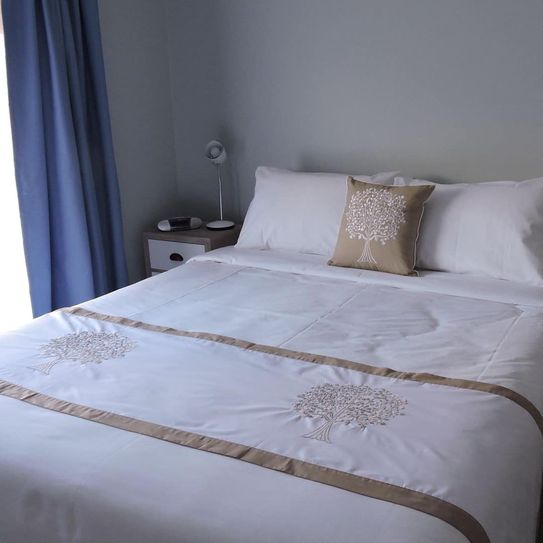 Bedroom with a king size bed, tan and white bedding, wooden bedside table, tall windows with blue curtains.