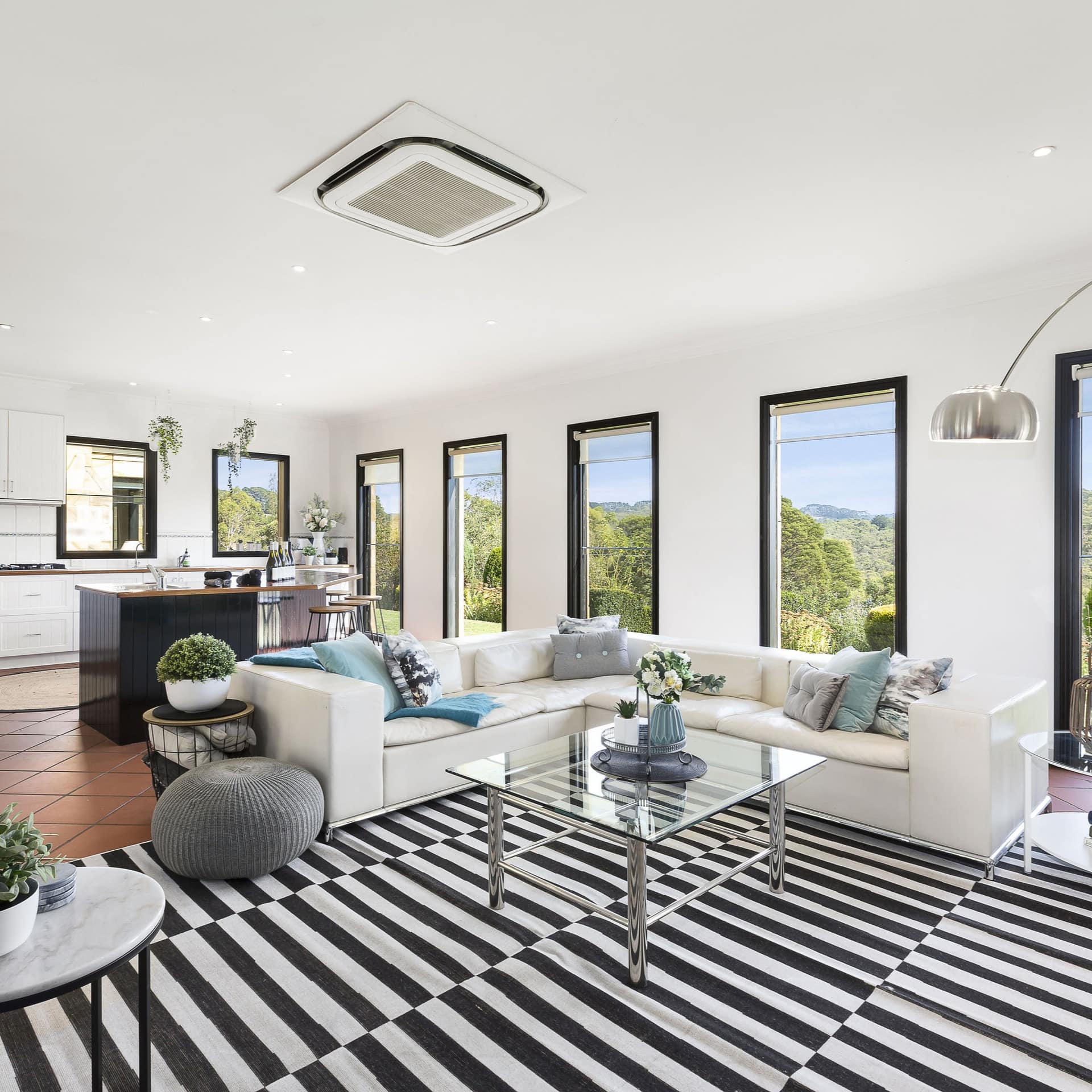 Open living room and kitchen with red tile floors, white sectional, glass table, black and white rug, and tall windows.