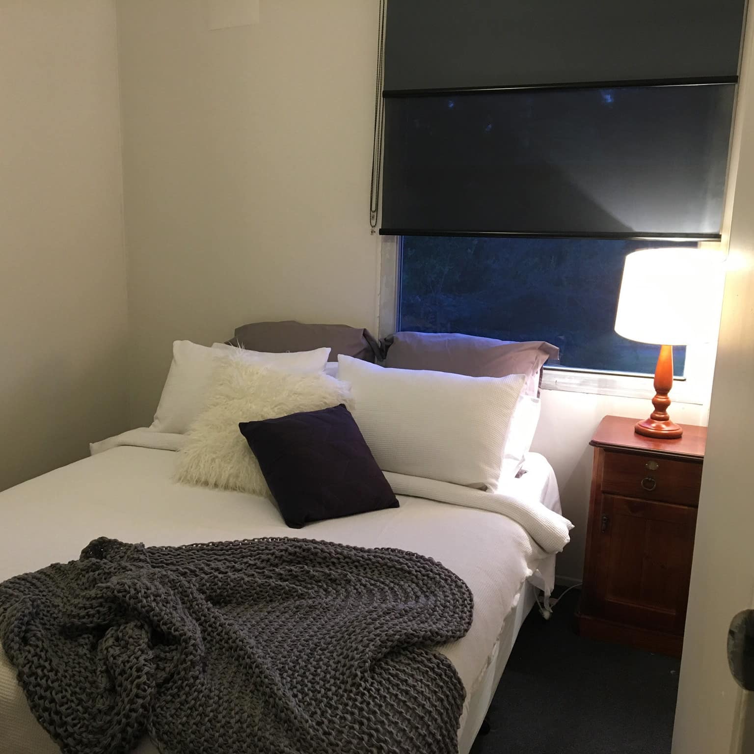 Bedroom with white walls, a king size bed, white and grey bedding, wooden nightstand, and tall window.