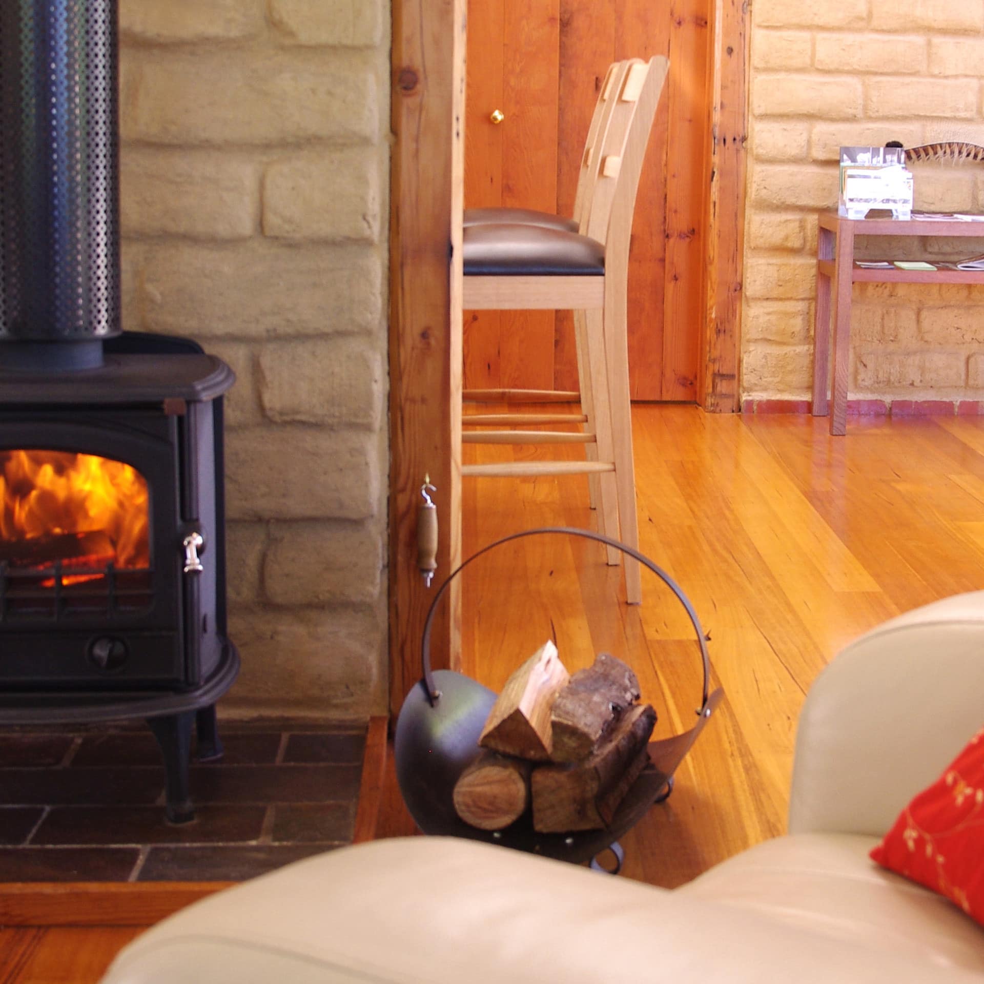 Open living room, hardwood floors, tan brick walls, a wood-burning stove in front of a beige leather chair with red pillow.