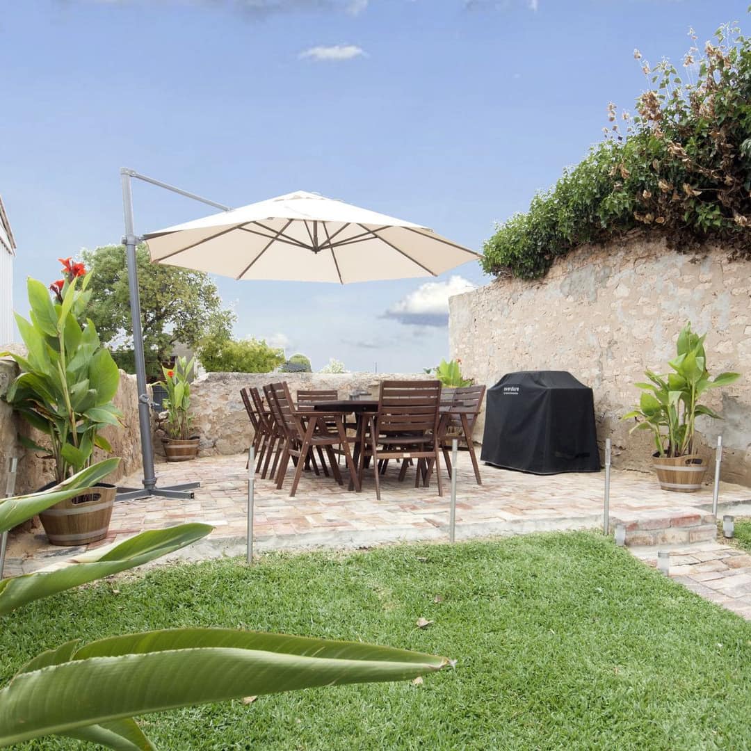 View of backyard enclosed by natural stone walls, stone patio with wooden dining set, umbrella, grill, and potted plants.