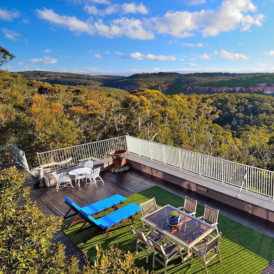 An expansive patio overlooking the Kanimbla Valley, white metal railing, glass dining table, and blue lounge chairs.