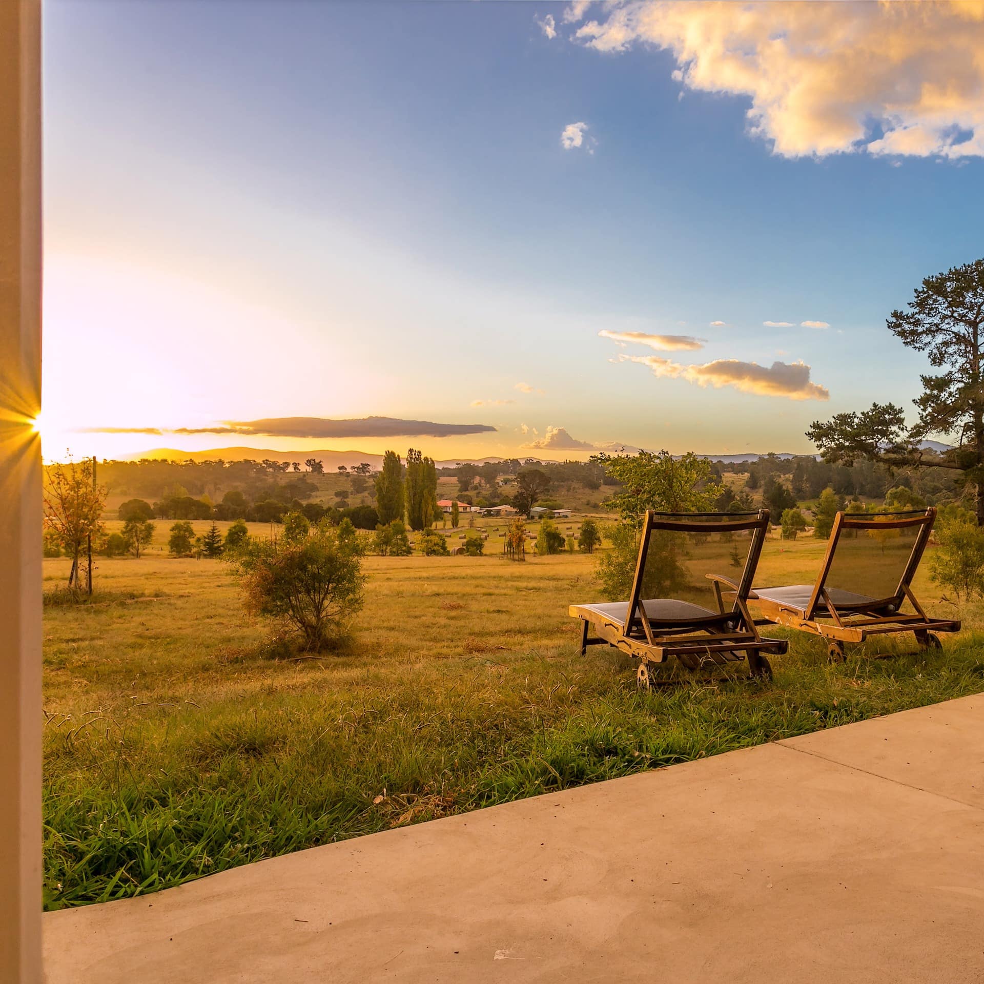 View of a picturesque sunrise over a rural, mountainous landscape from a patio space with wooden lounge chairs. 