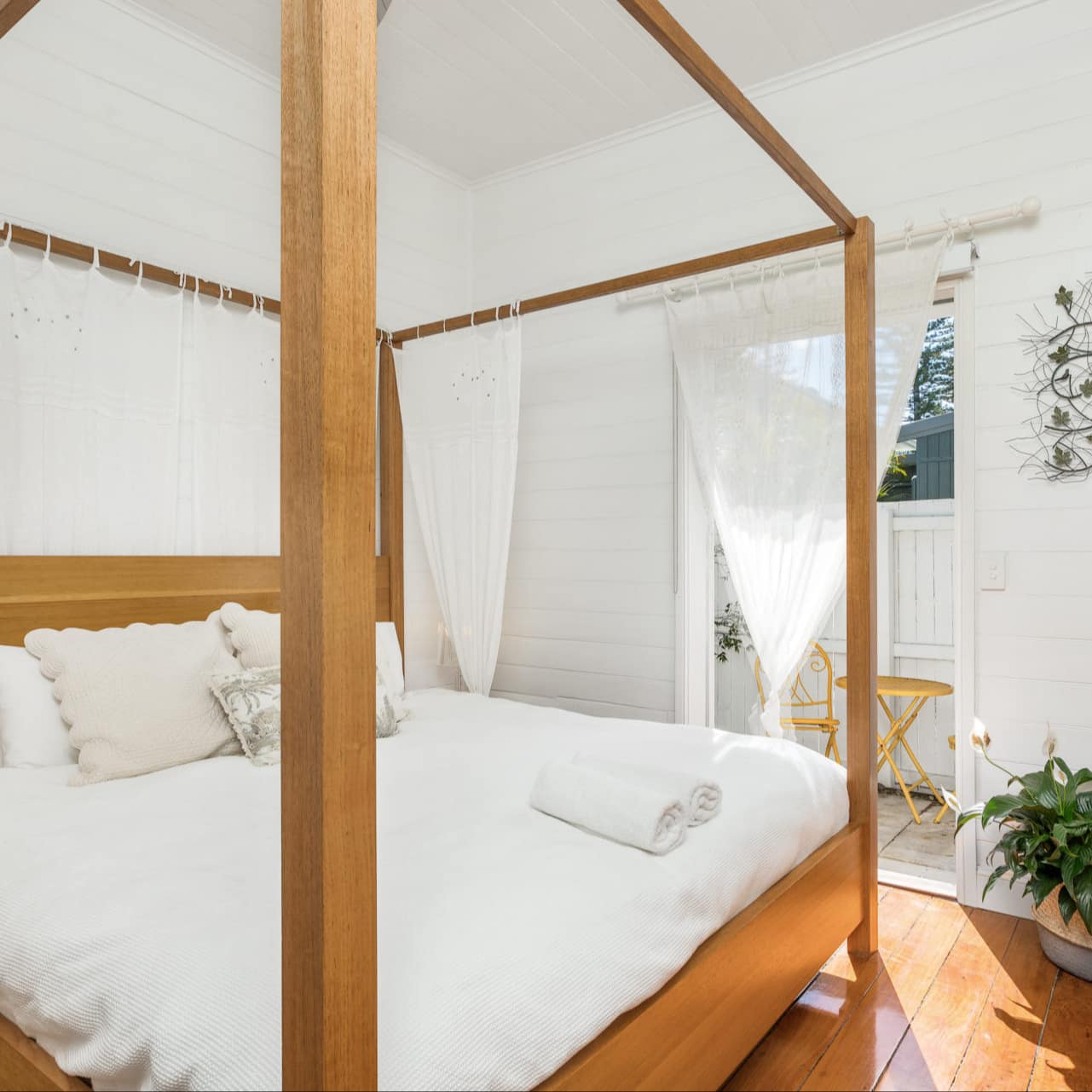 Sunny bedroom with hardwood floors, white walls and a wooden canopy bed with white bedding and sheer canopy.