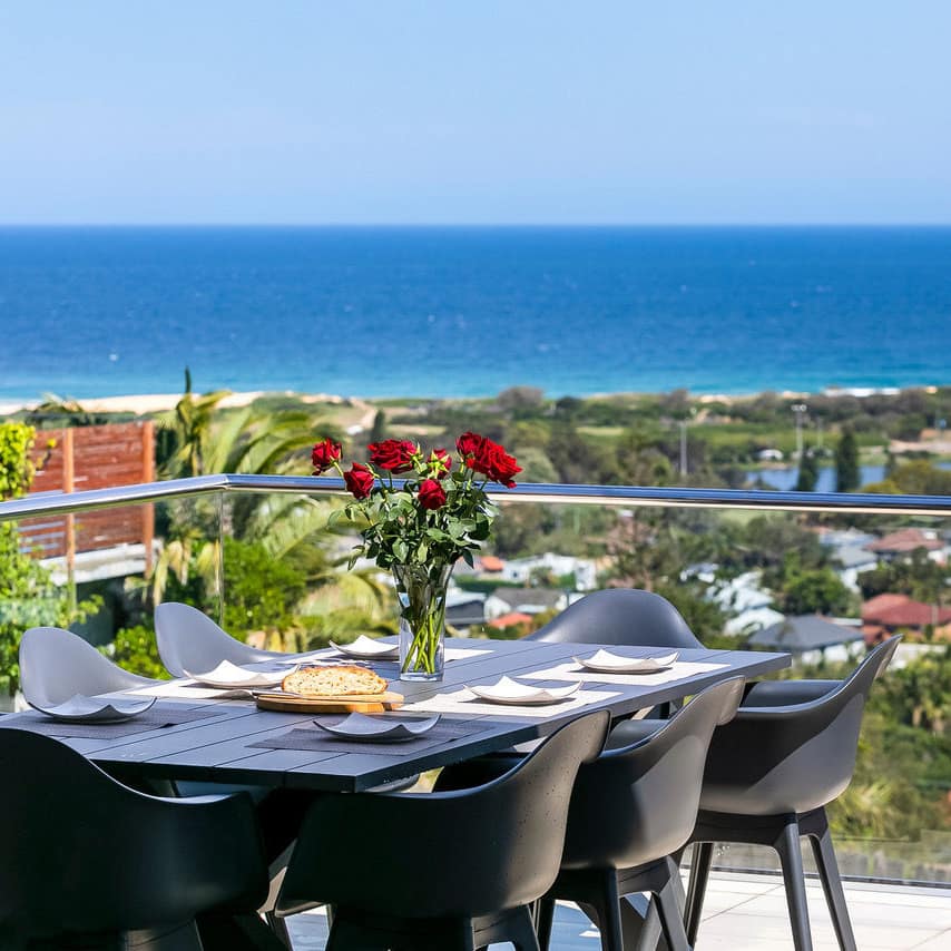 180 degree panoramic view of town and ocean from glass-enclosed terrace with outdoor dining table with vase of roses