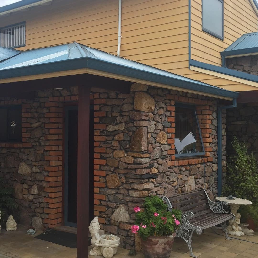 A 2-storey house with a stone and wood exterior painted in gold with a green metal roof, a patio and potted plants.