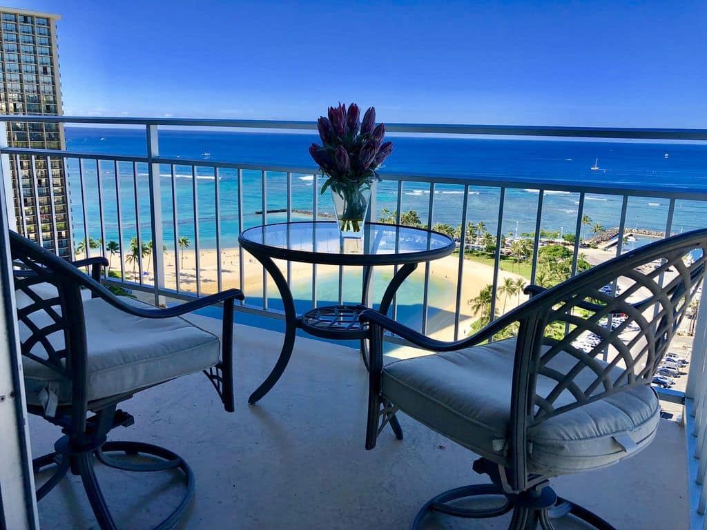 Two chairs and a table on balcony of condo overlooking the beach full of palm trees and aqua water