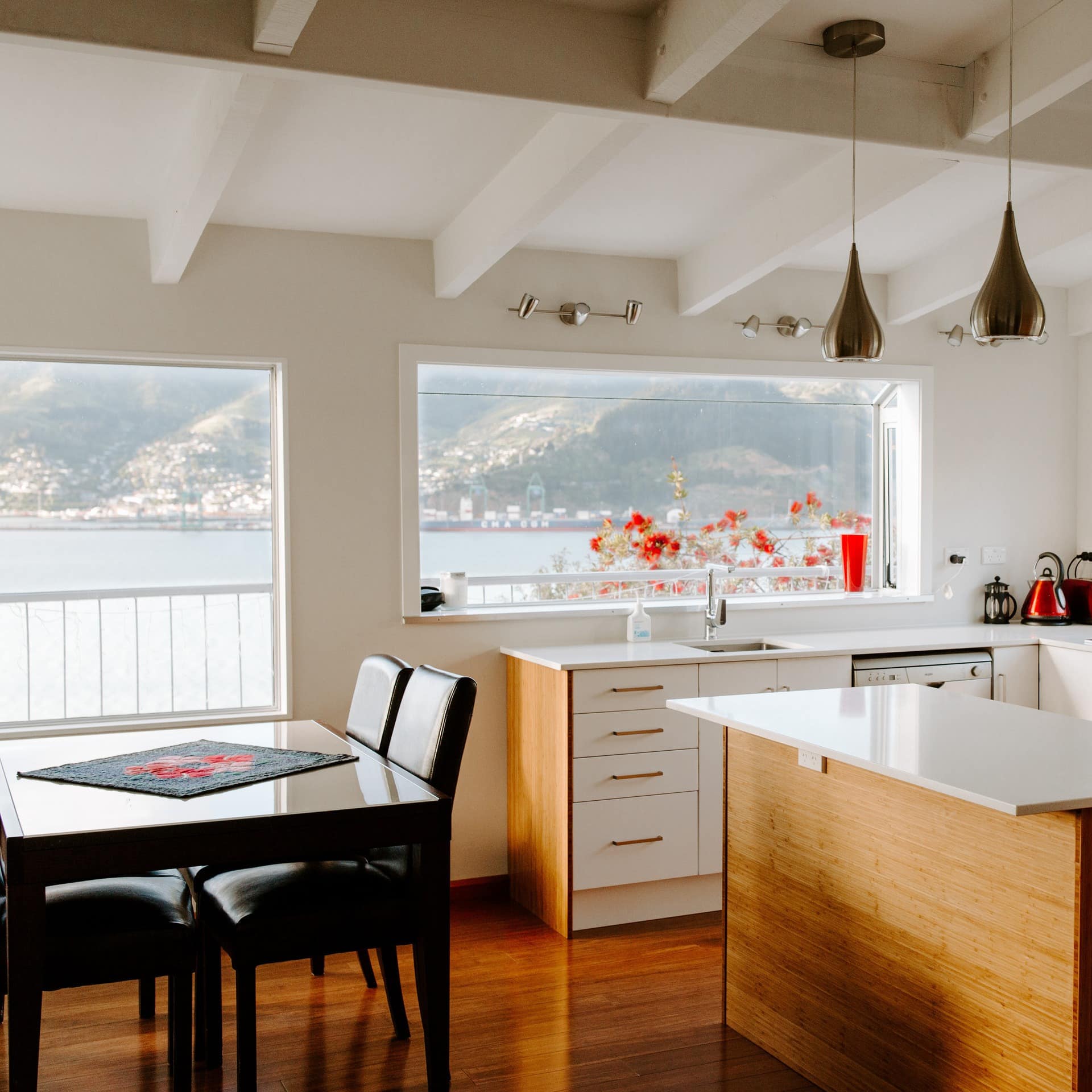 A modern kitchen and dining room with white cabinets and countertops, a black dining set, wide windows with harbour views.