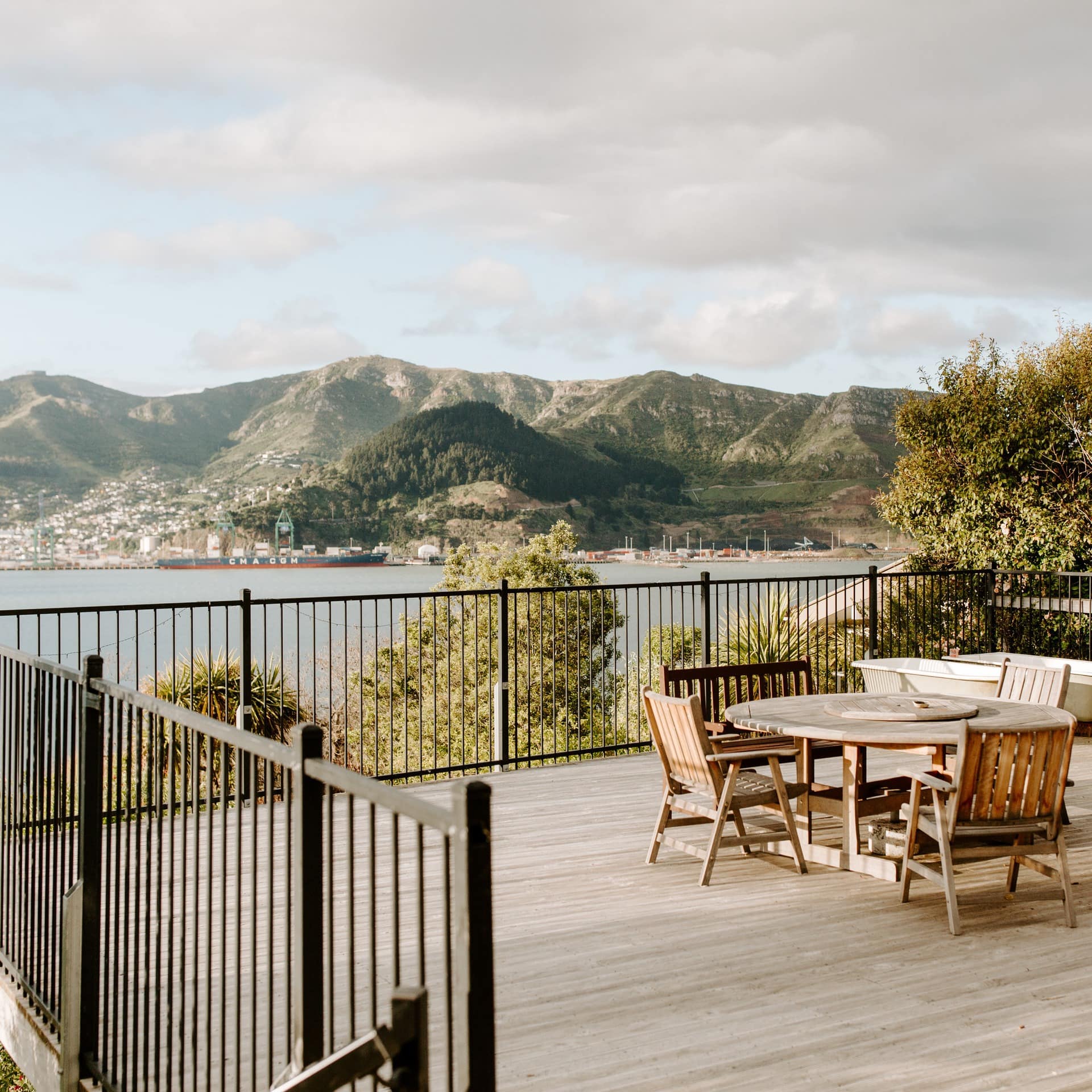 A wooden deck with black metal railings, wooden dining set, outdoor tubs, overlooking the harbour and mountains.