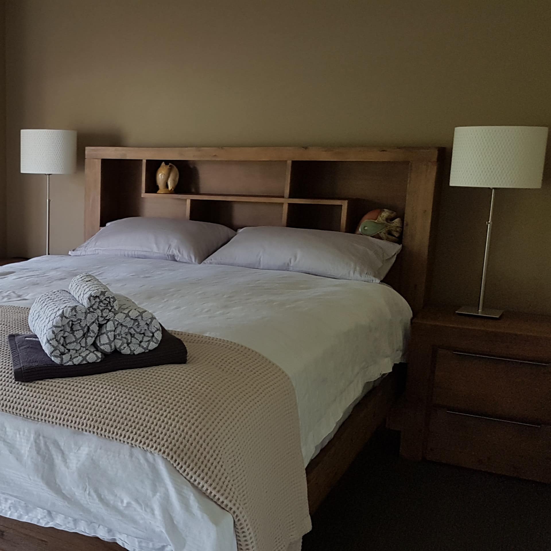 Bedroom furnished in neutral tones, king size bed with wood bed frame, white linen, tall windows with tropical plant views.