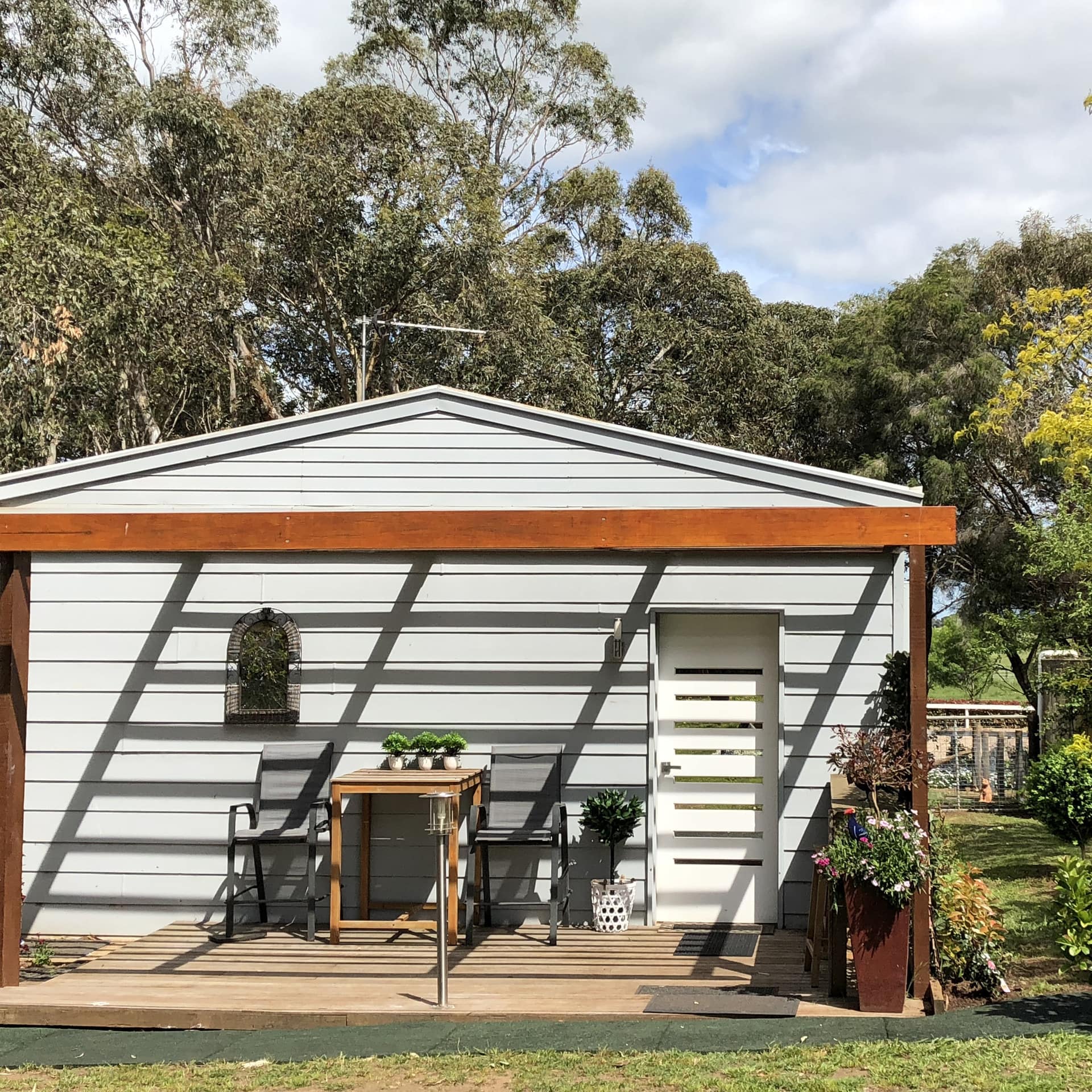 A modern cottage with grey siding, wooden porch, grey lounge chair, wooden table, surrounded by lush greenery.