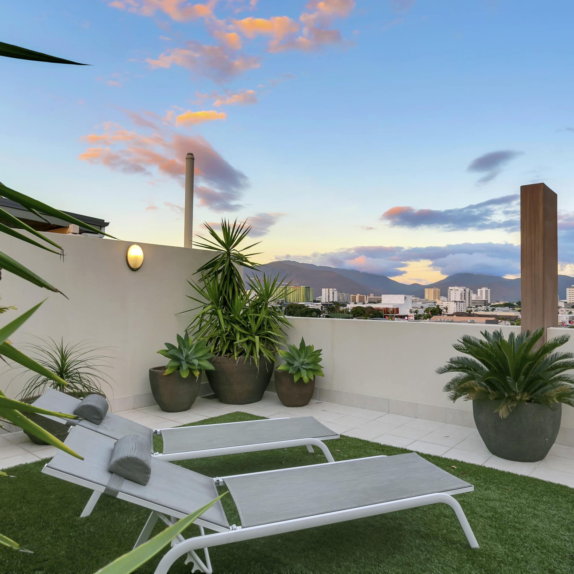Two grey lounge chairs sits on a patch of artificial grass on a white tile balcony overlooking the town of Cairns.