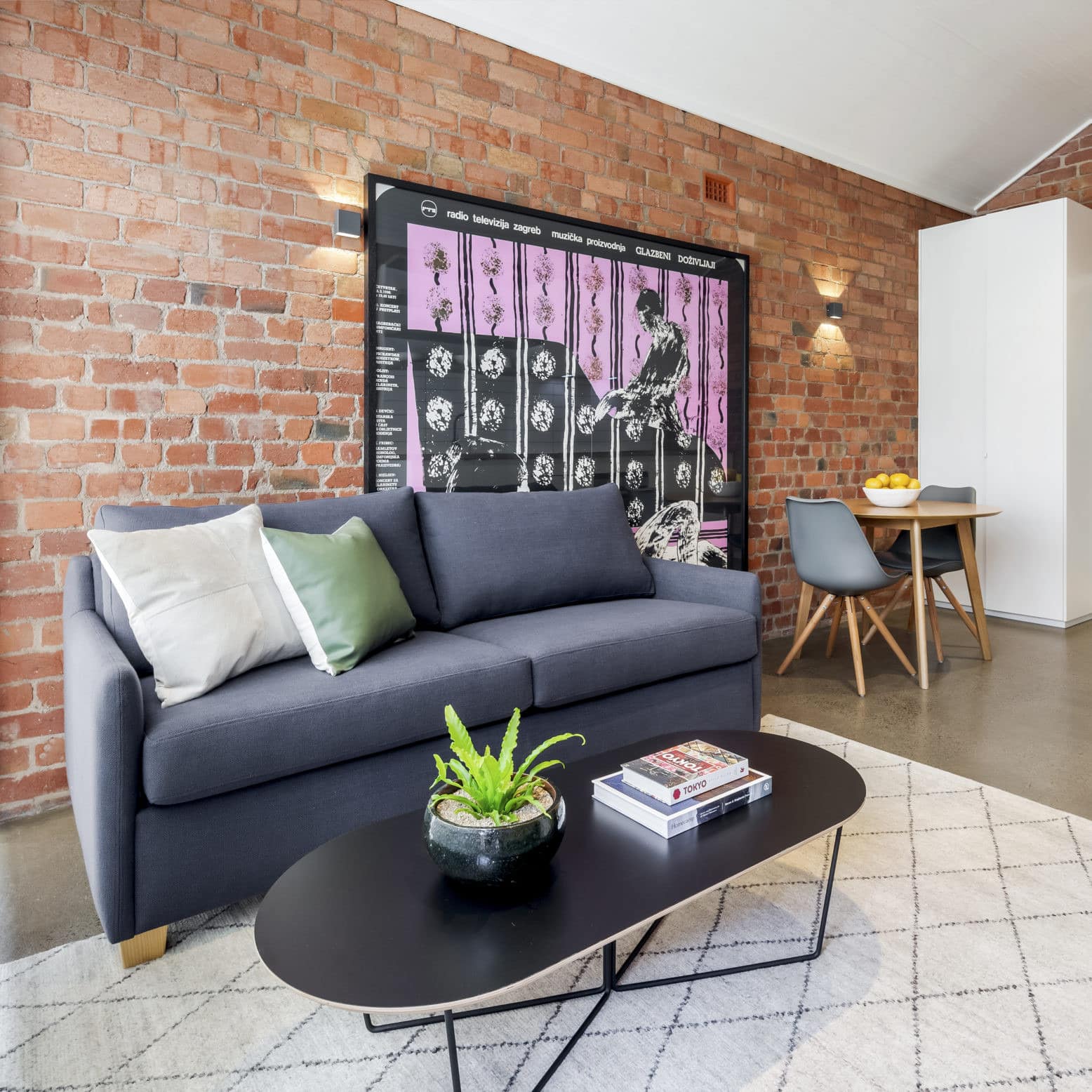 Modern dining and living room with navy blue soda, grey stone floors, exposed brick walls, and a wooden dining set.