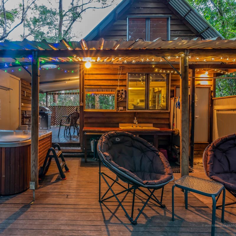 View of a wooden deck of a wood cabin with a hot tub, round black chairs, and string lights on the porch covering.
