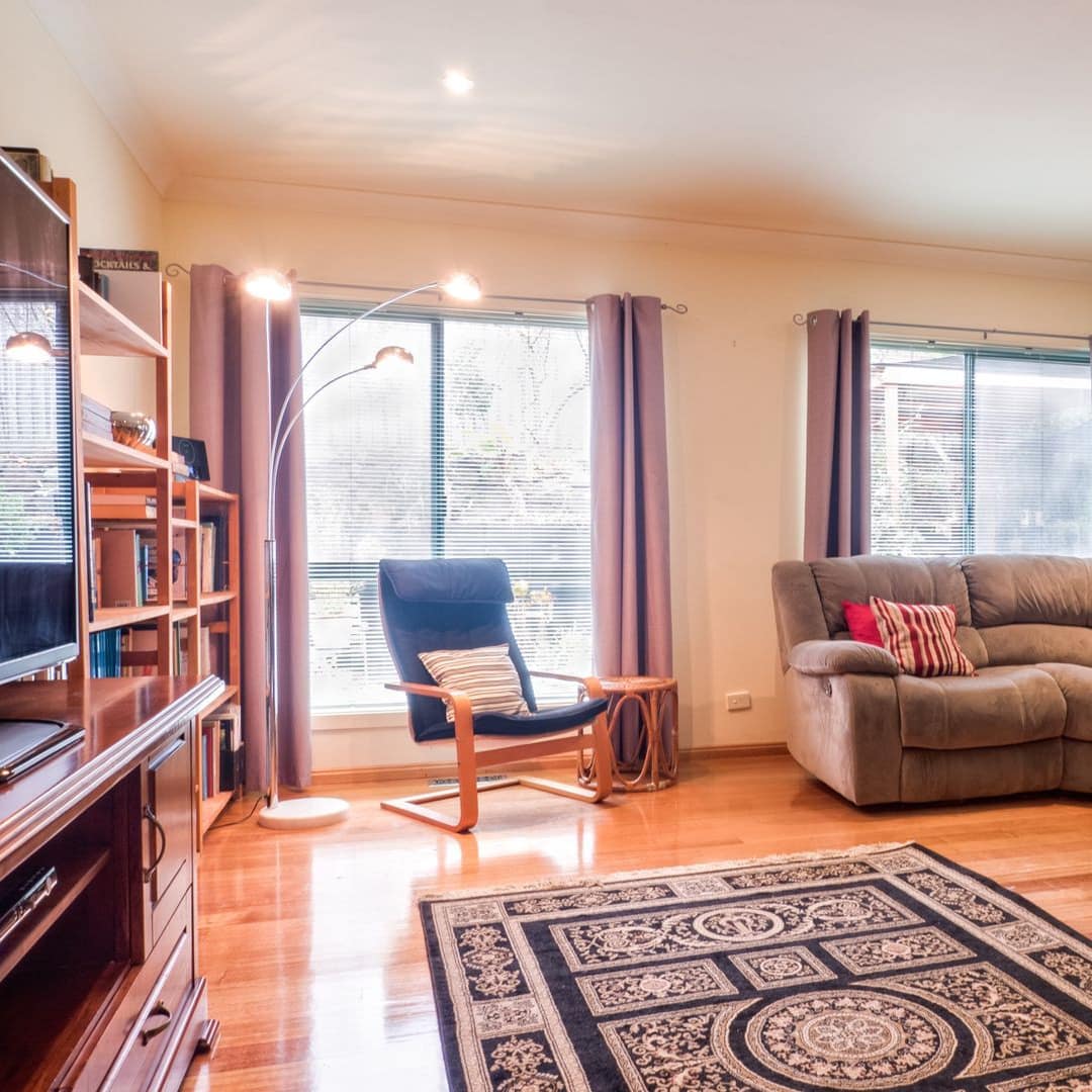 Open living room with hardwood floors, brown suede sectional, flatscreen TV, and tall windows with red curtains.