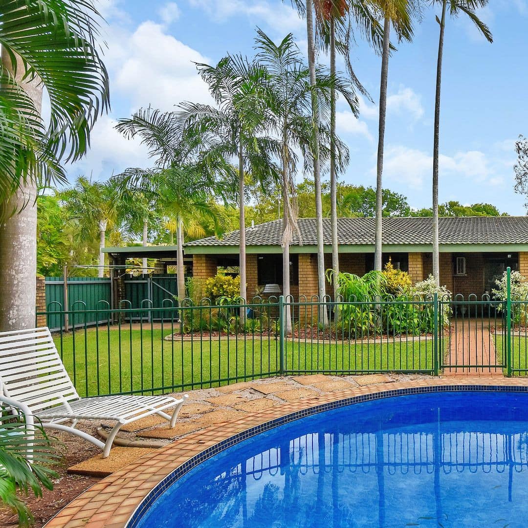 Exterior view of ranch-style home with private curved pool enclosed by green iron fence, and lush lawn with palm trees.
