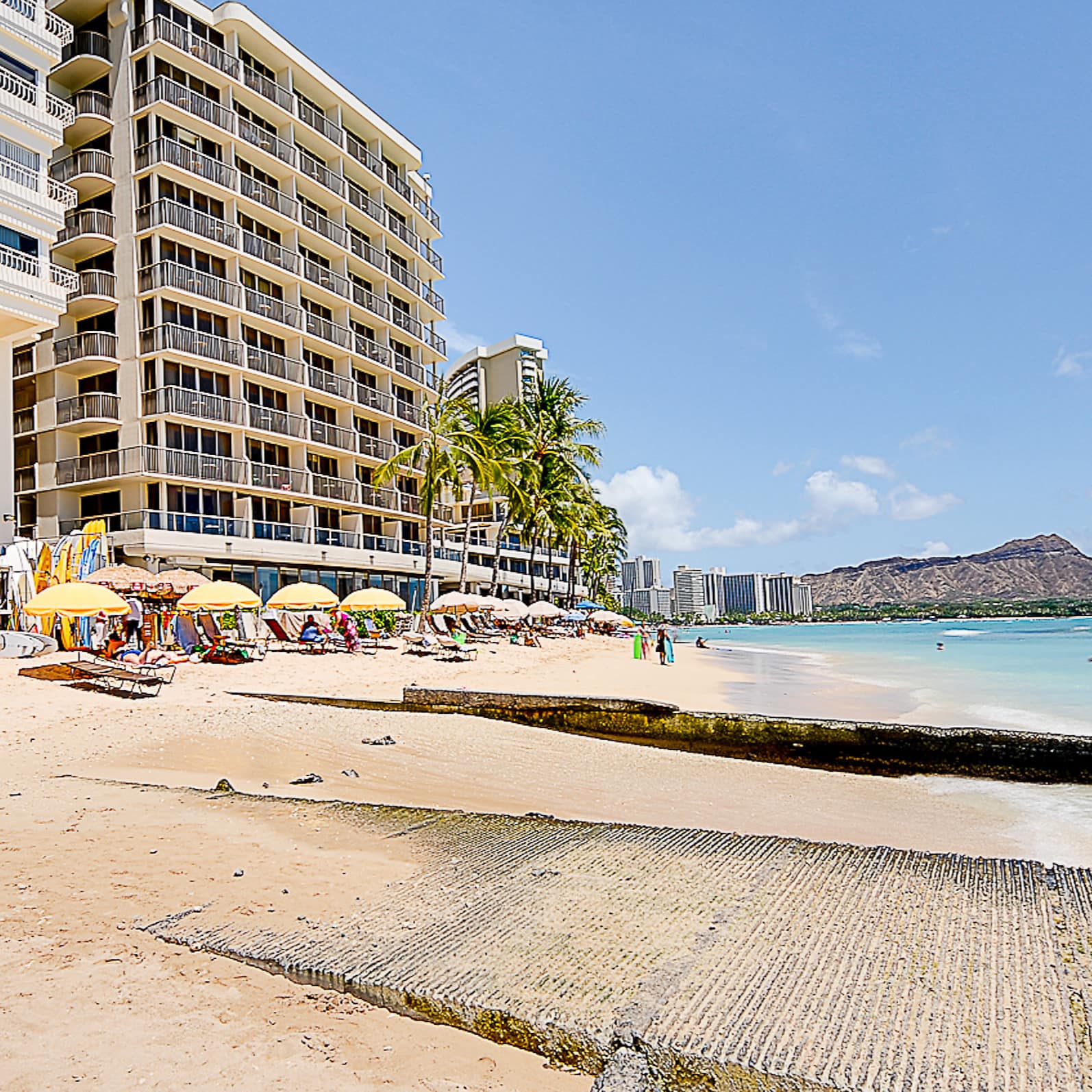 Large apartment building in front of ocean, palms, and yellow umbrellas on the beach with mountain in the background
