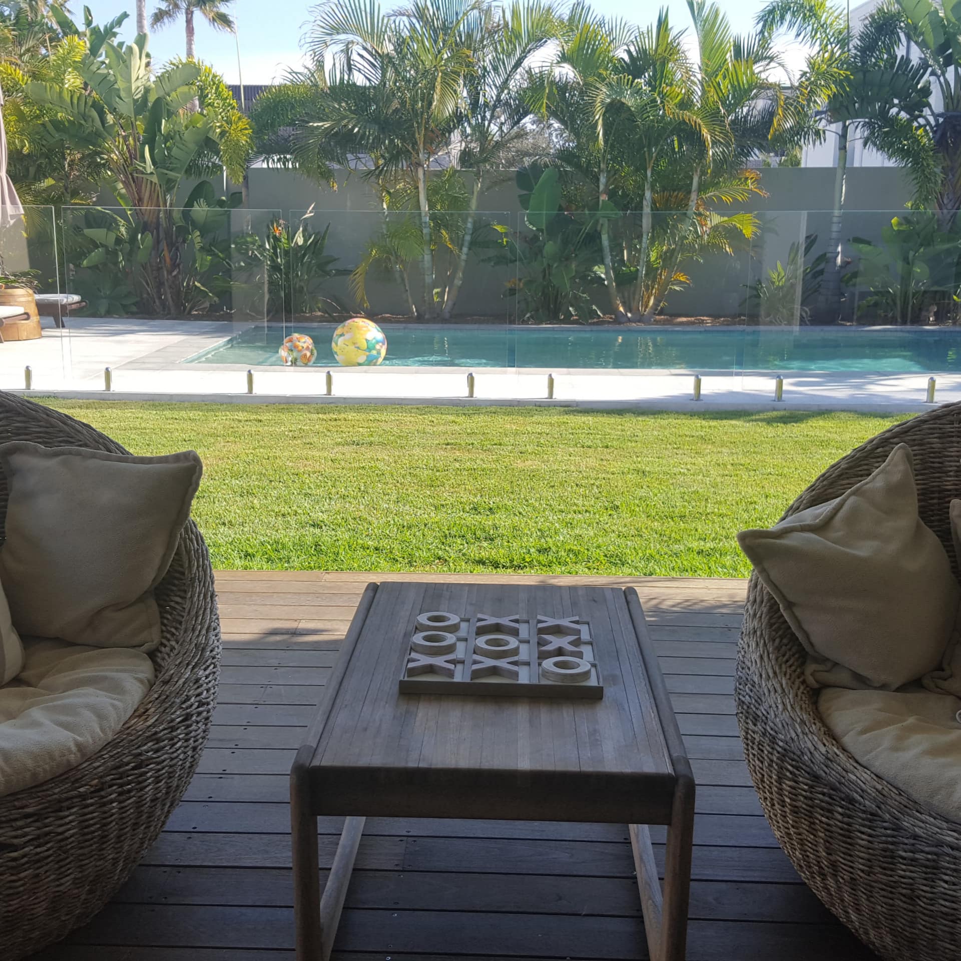 Two wicker rattan ball chairs on wooden deck sit in front of a sunny lawn and private pool, palm trees surround the pool.