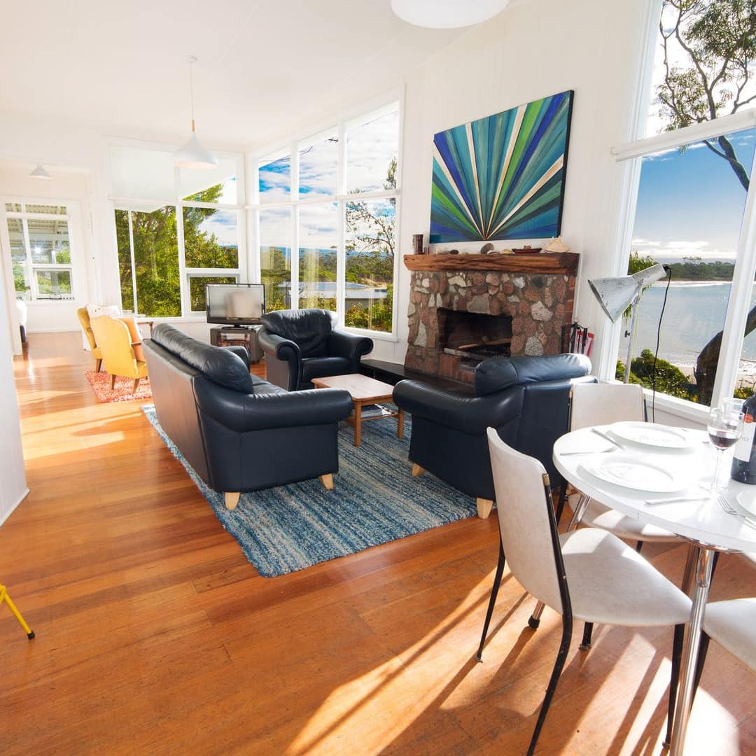 Open living and dining room, white dining set, black leather sofa and chairs, natural stone fireplace, tall windows.