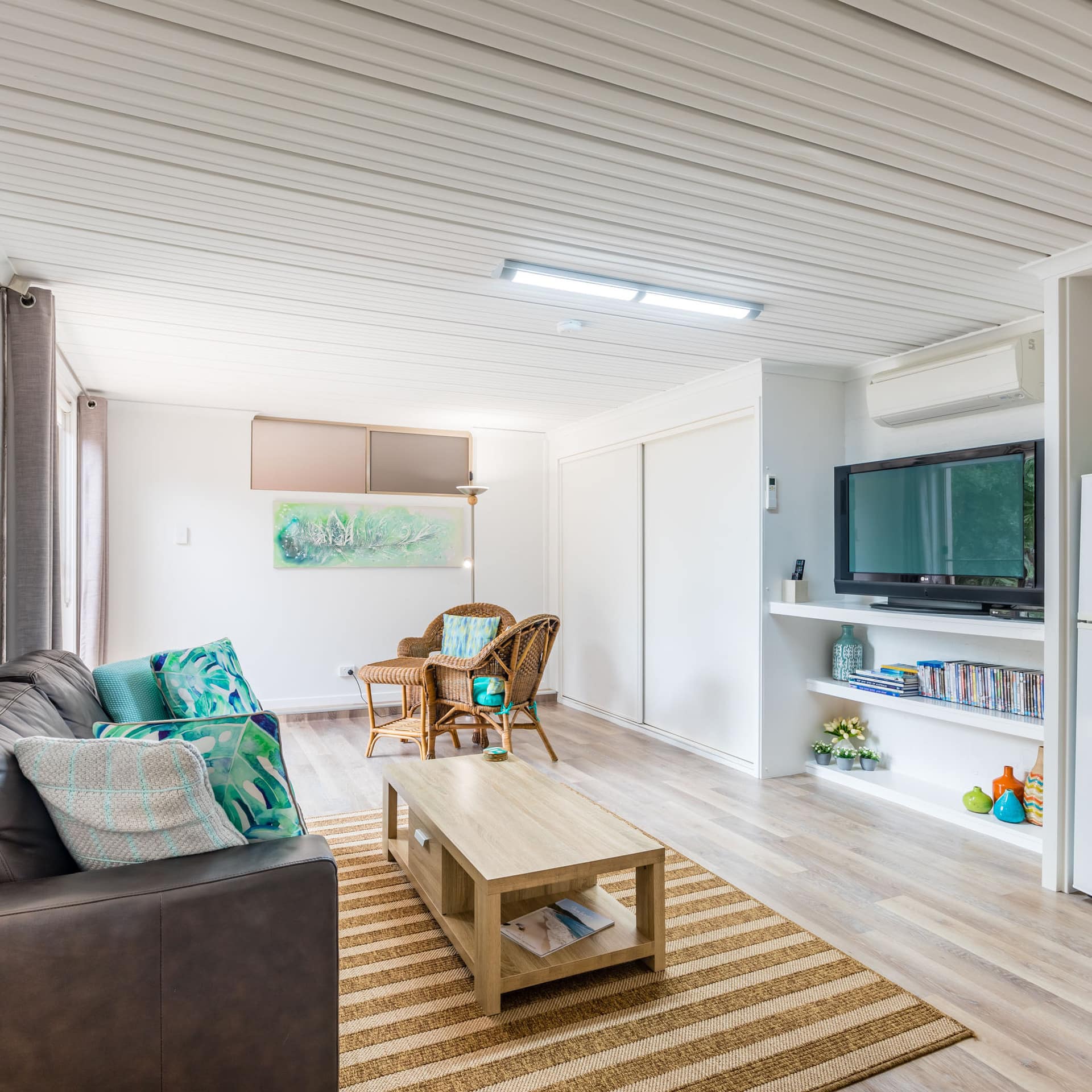 Open living room with hardwood floors, white walls, brown leather sofa, wicker dining set, and flatscreen TV.
