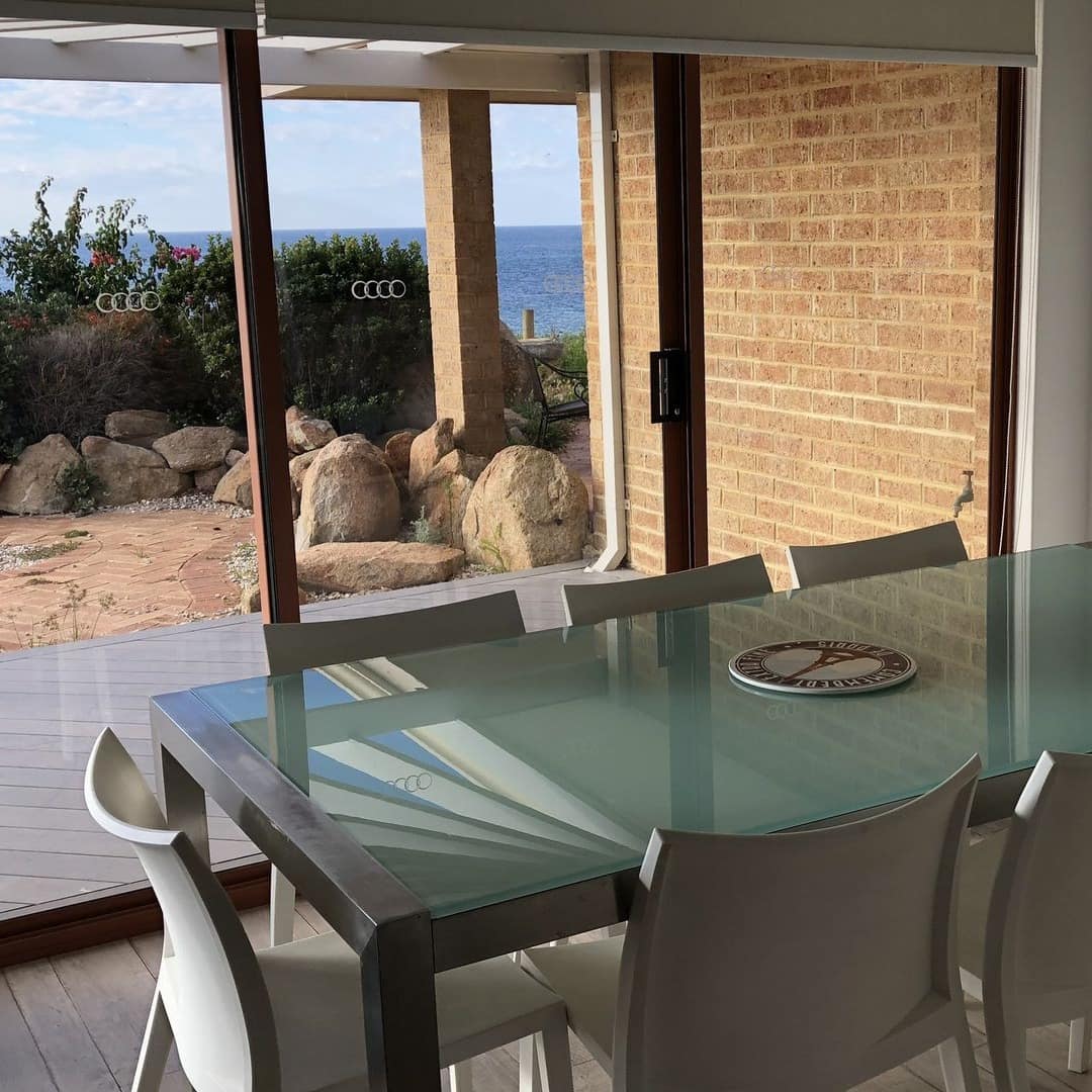 Dining room with a glass-top modern table and chairs and sliding glass doors opening to a wooden deck and beachfront.