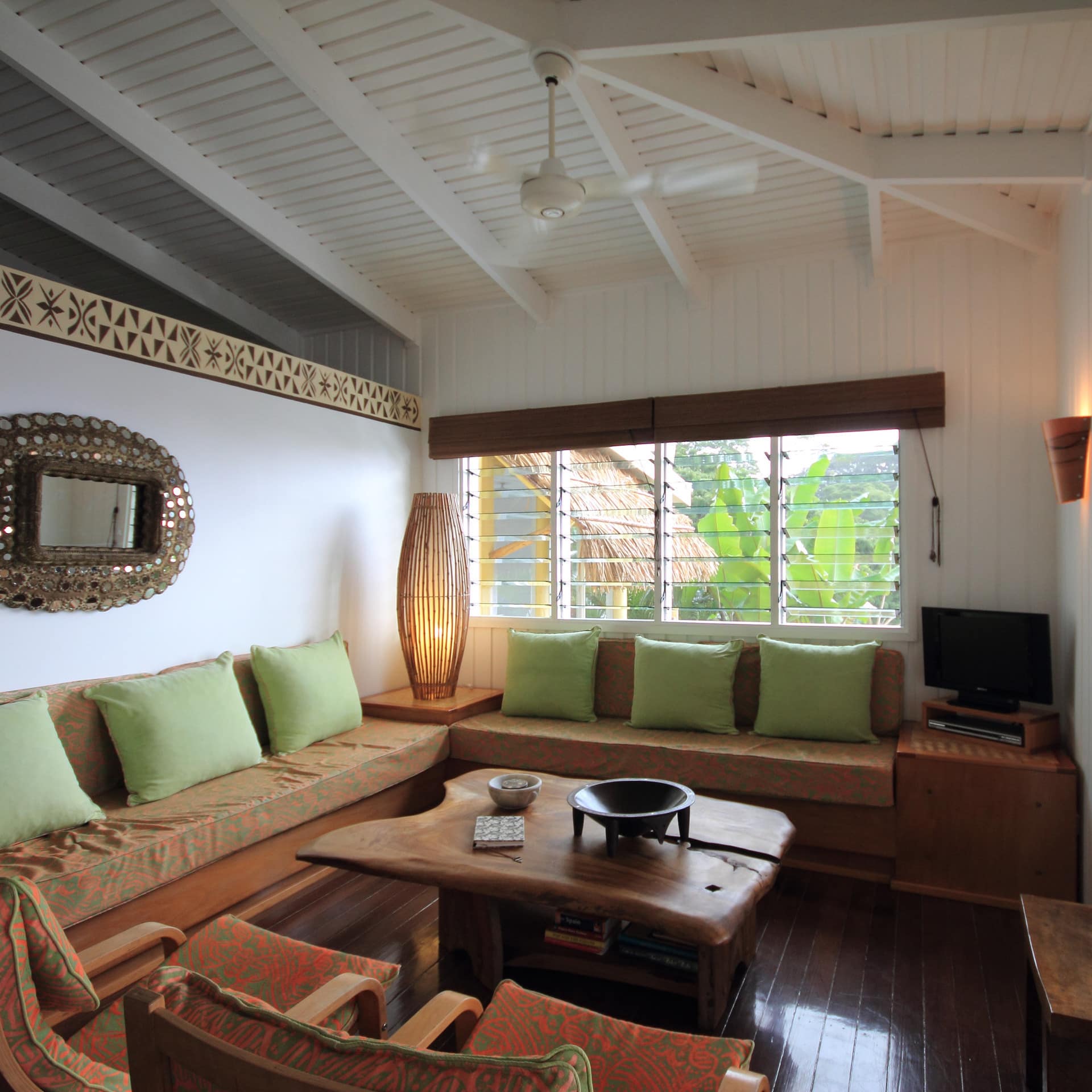 Living room with hardwood floor, white panelled walls, brown sofa, natural wood table, and wide windows.