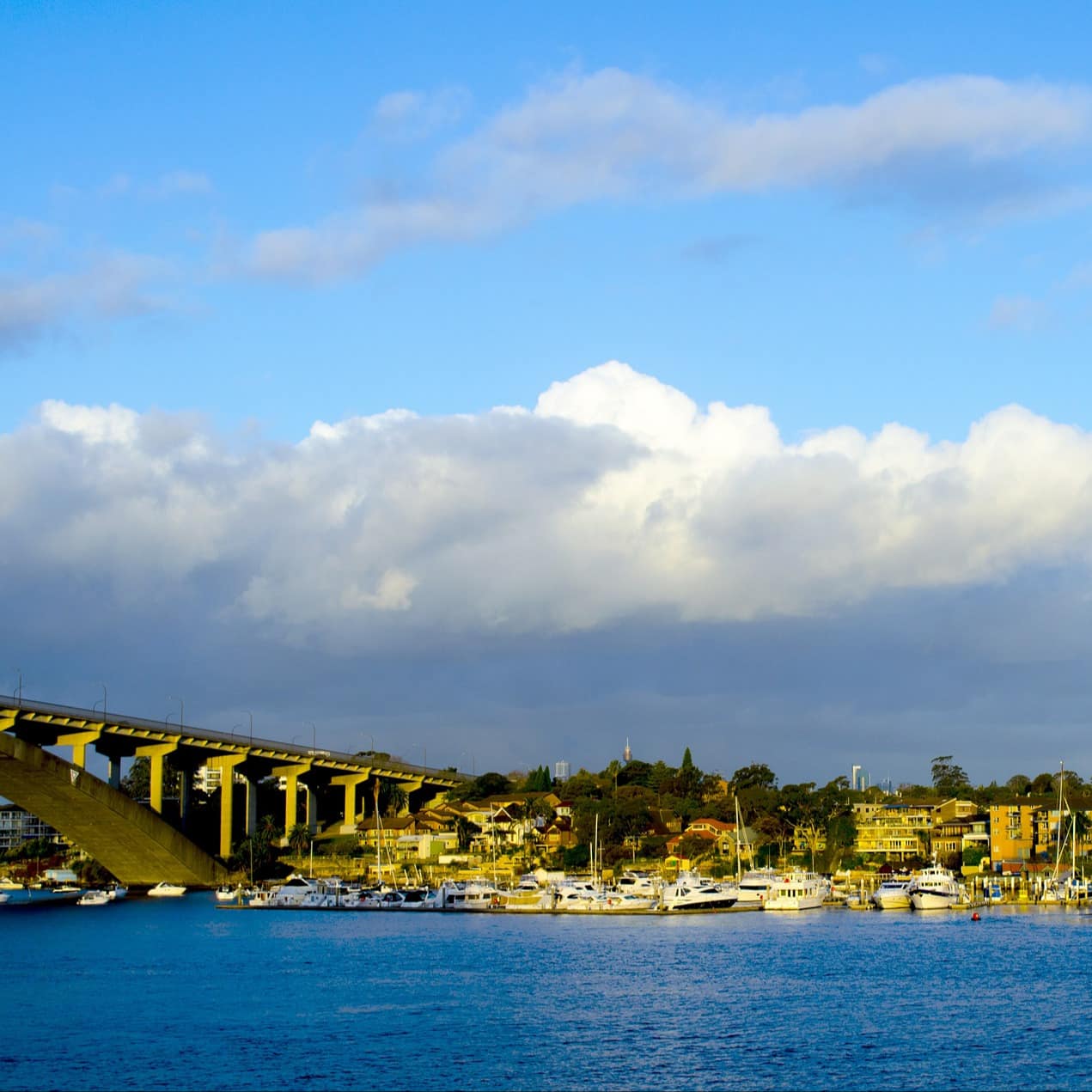 Gladesville Bridge in Parramatta with royal blue water, boats at harbour, and the town visible beyond with a bright blue sky