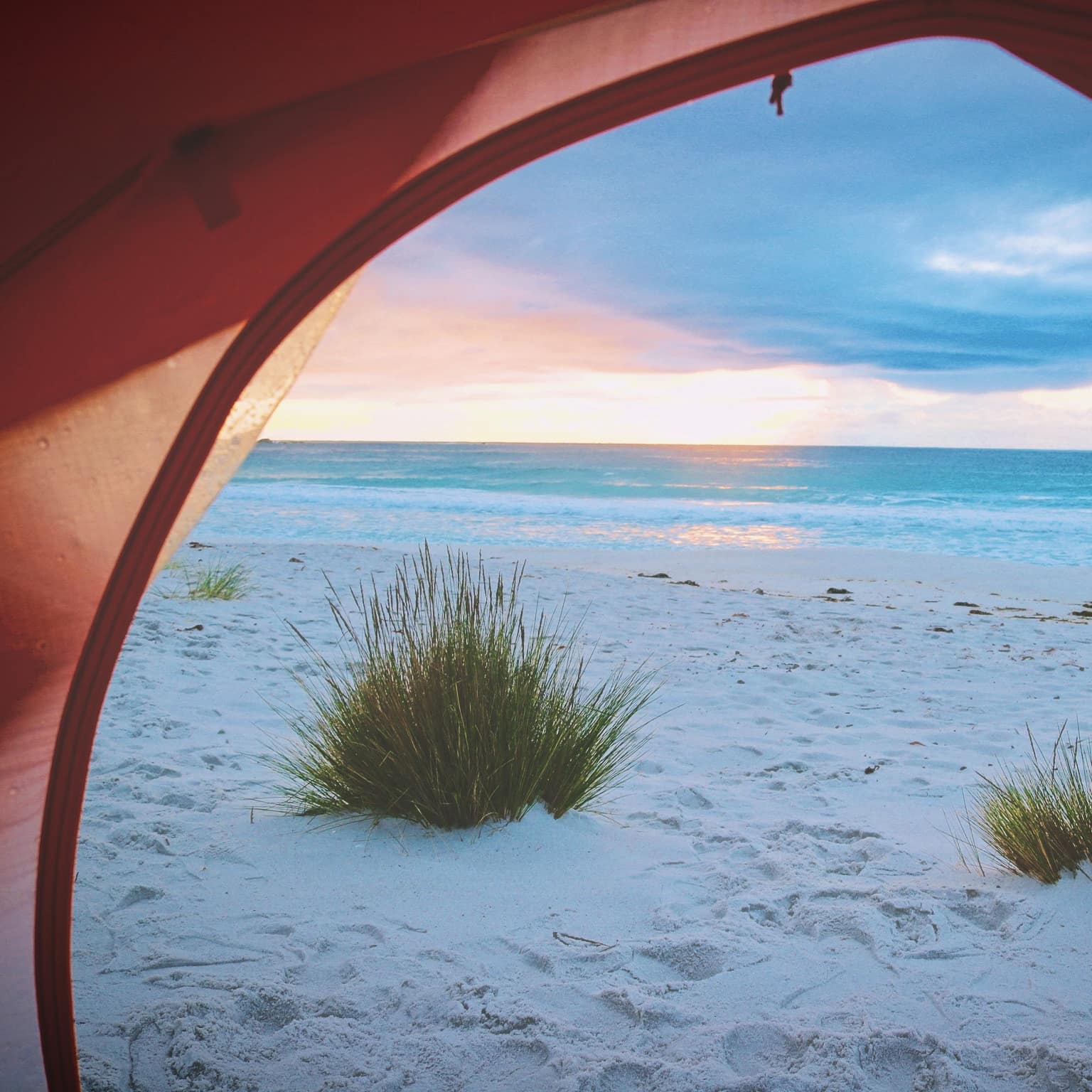 A view of the sunrise on the ocean, soft sands and sea grass through a rounded tent opening