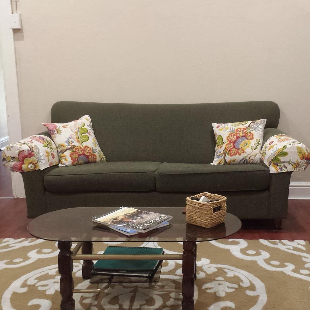 Living room with hardwood floors and beige walls, dark green canvas sofa, floral throw pillows, tan and white area rug.