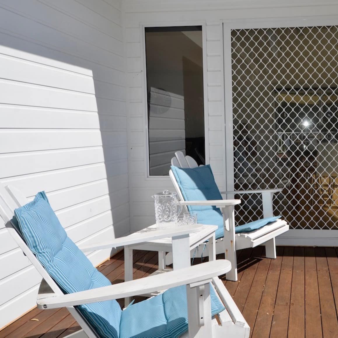 Hardwood deck of home with white vinyl siding, white lounge chairs with blue cushions, and open sliding glass door.
