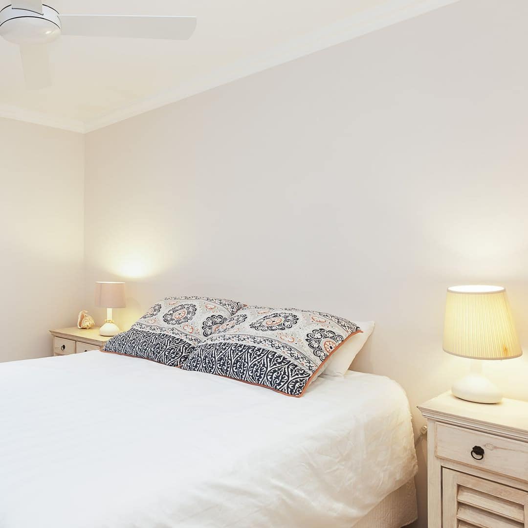 Bedroom with white walls and carpet, tall windows with ocean views, a queen size bed, white linen, and tan curtains.