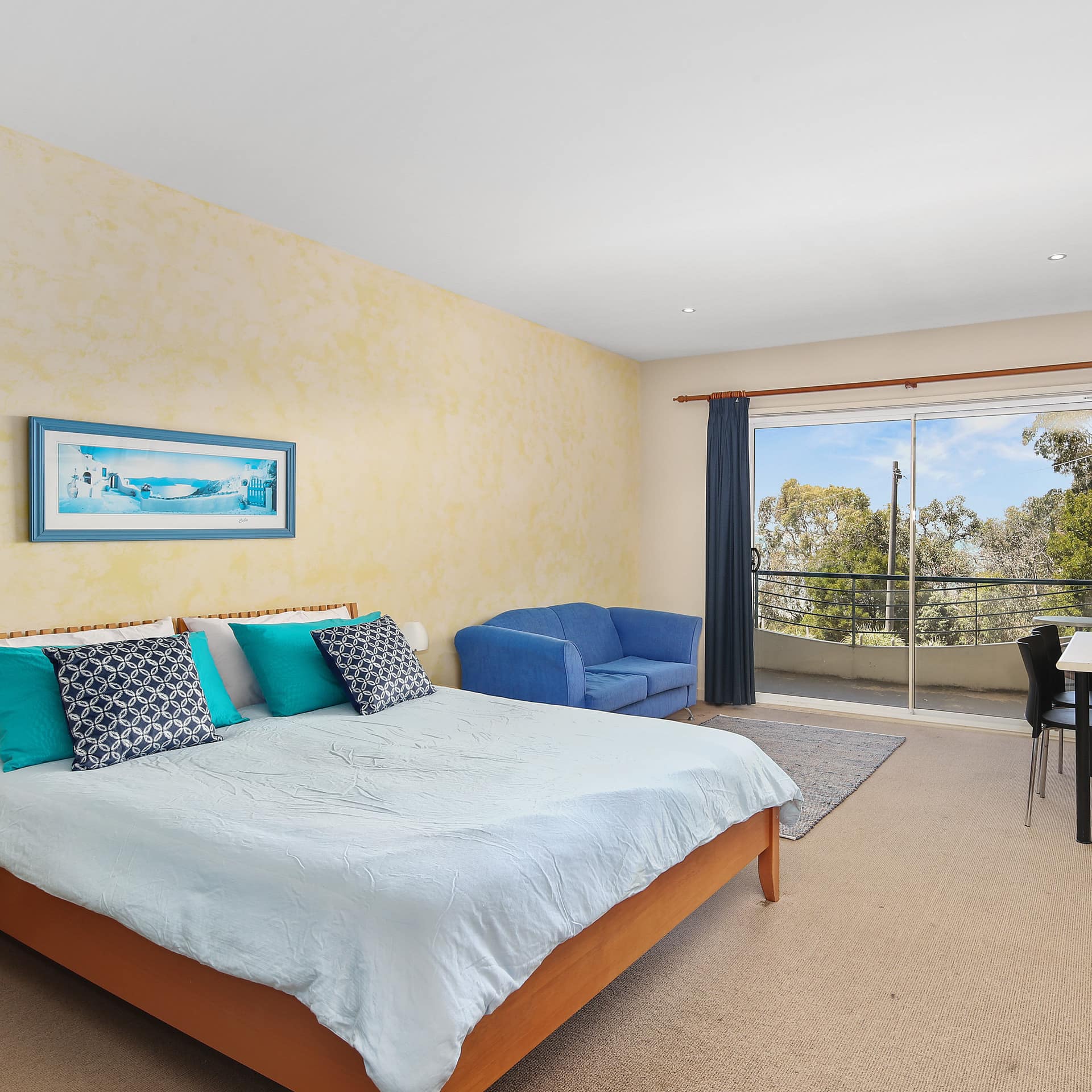 Spacious bedroom with beige carpet, yellow walls, a king size bed with blue bedding, attached balcony.