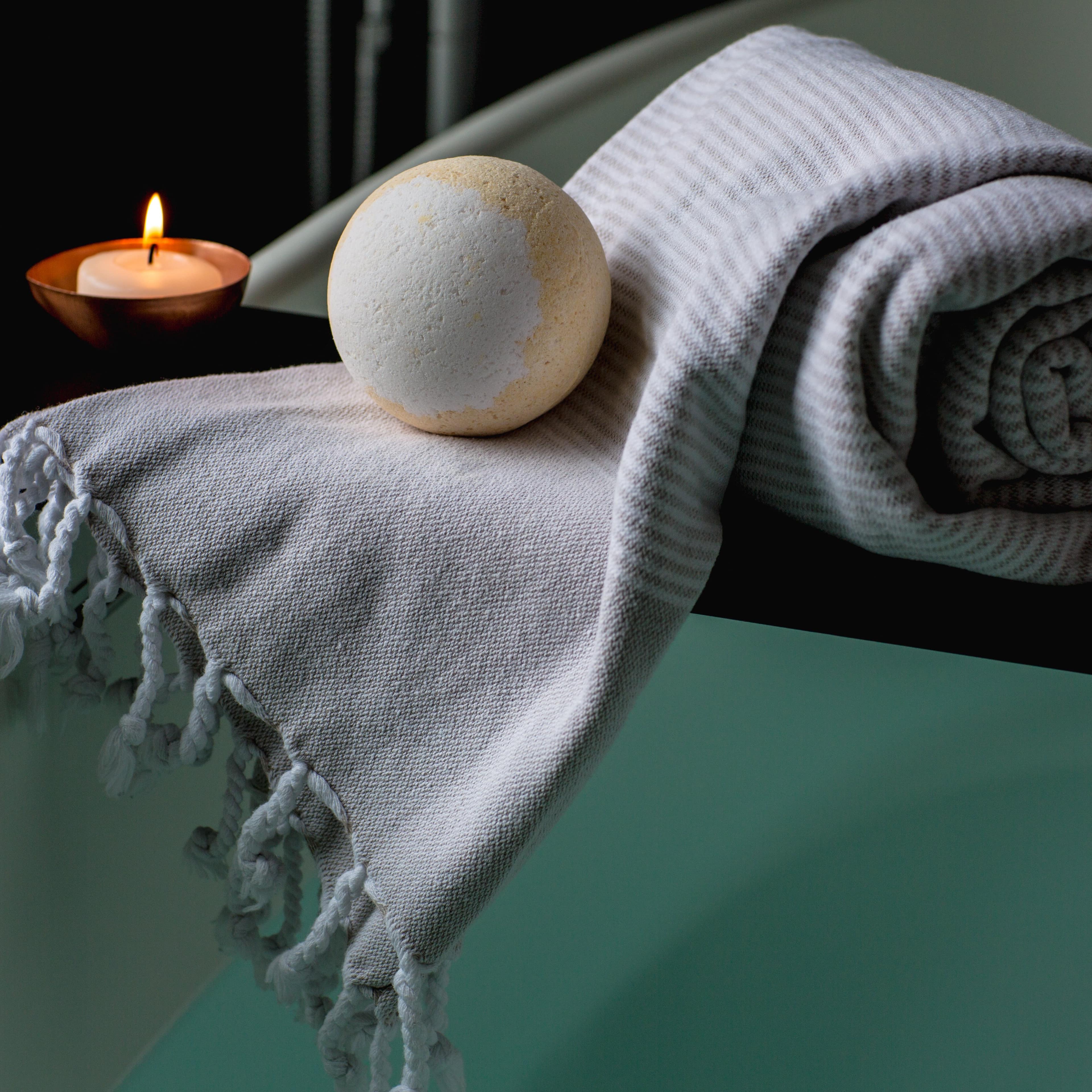 Towel, bath bomb and lit candle sit on wooden shelf above spa tub