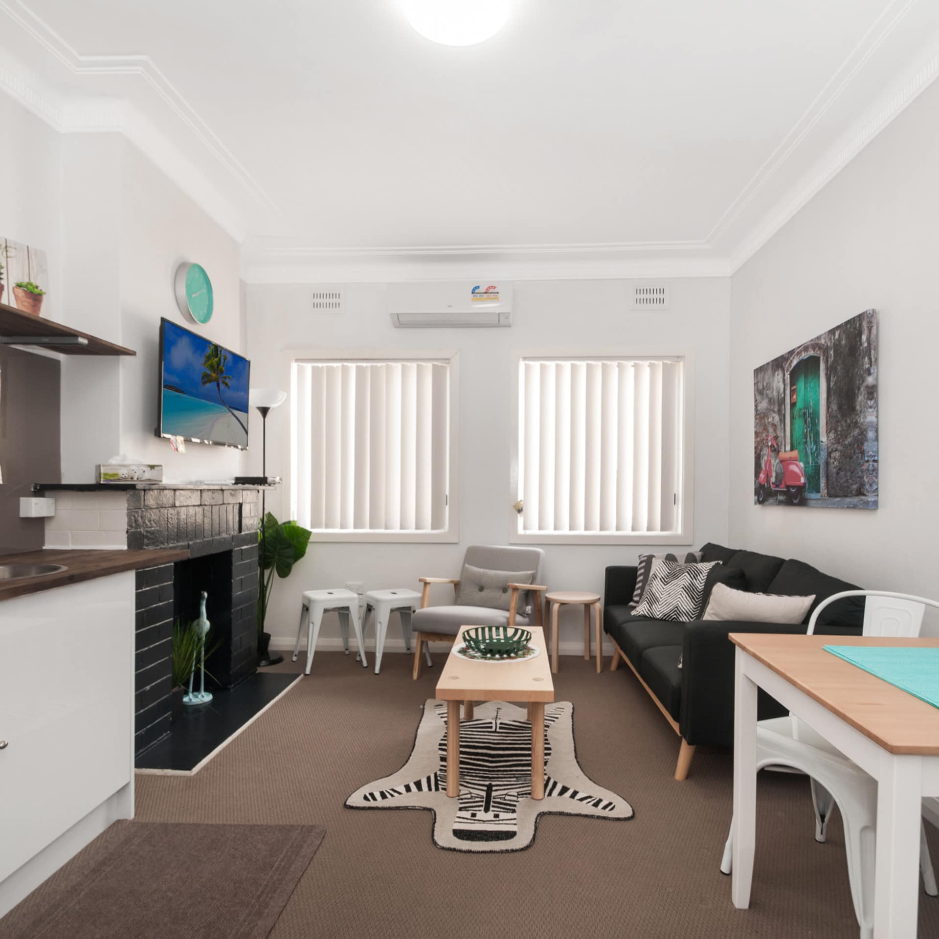 Open kitchen and living room with tan carpet, white walls, white kitchen cabinets, black brick fireplace, and modern decor.
