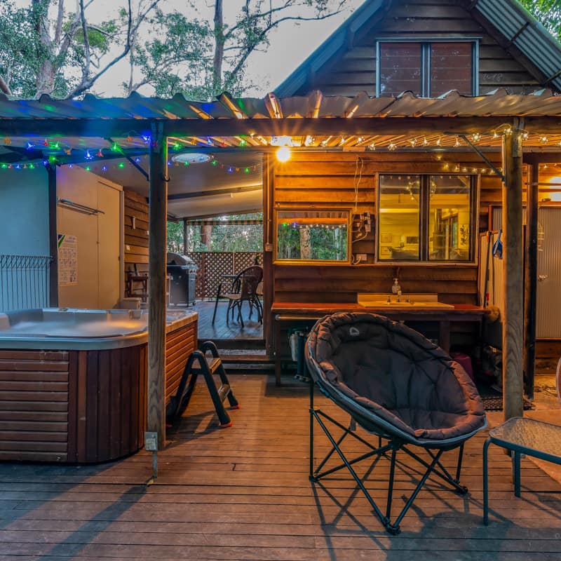 Evening view of the covered deck of the wood cabin with a hot tub, round lounge chairs, colorful lights line roof.